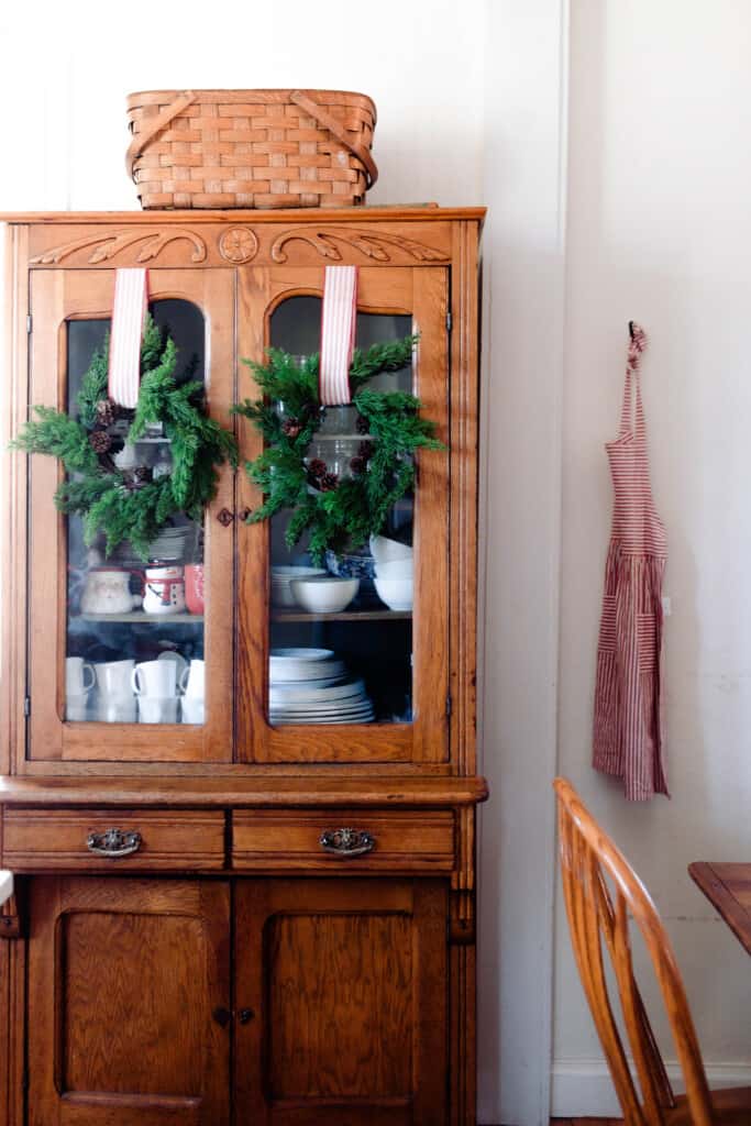 vintage hutch with two greenery wreaths with pinecones hang from a white and red ticking stripe ribbon. a red and white striped apron hang on the wall to the right.