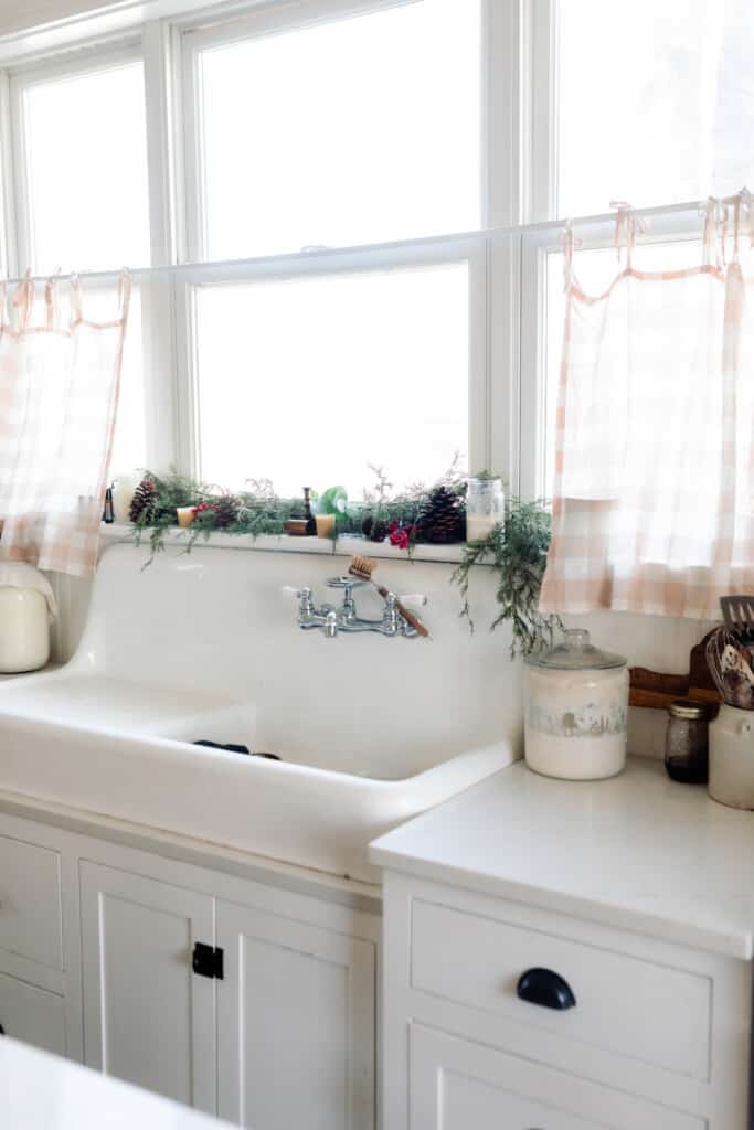 vintage farmhouse sink with a window above. The windowsill is decorated with pine cones, bells, candles, and greenery.