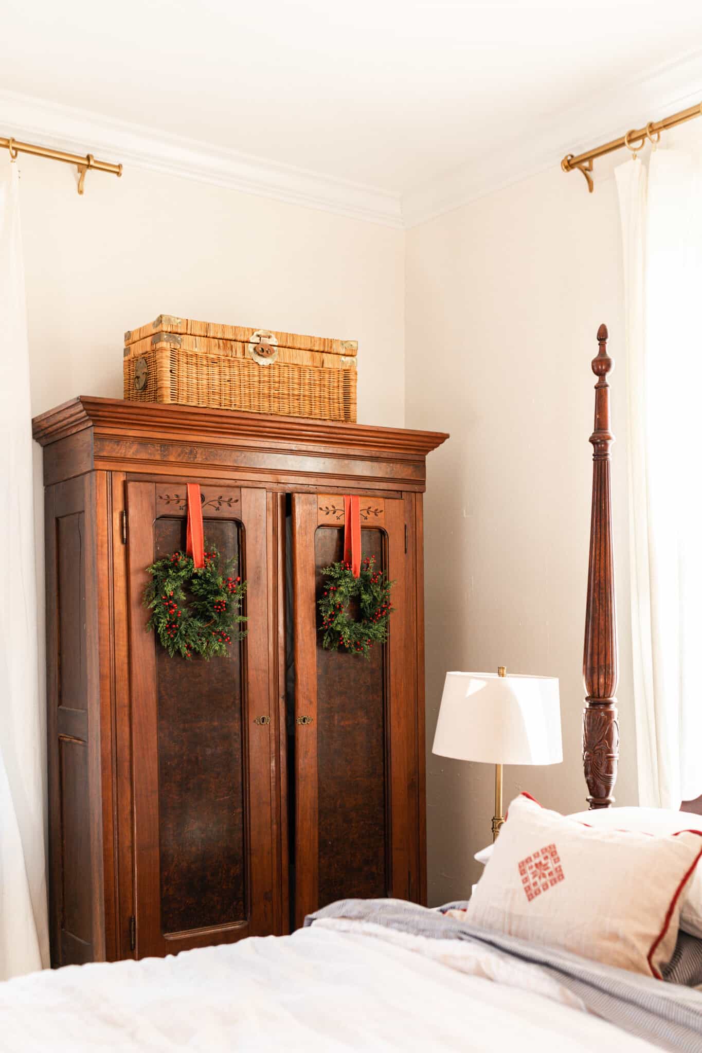 a corner of a vintage four poster bed decorated with white linens, and a cream and red holiday inspired pillow. A large armoire with wreaths hanging by red ribbon is next to the bed.