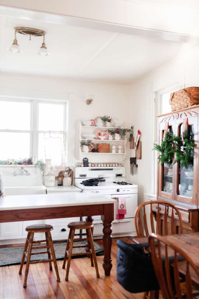 a white farmhouse kitchen with pops of red throughout the space with greenery. Wreaths hang from an amour. 