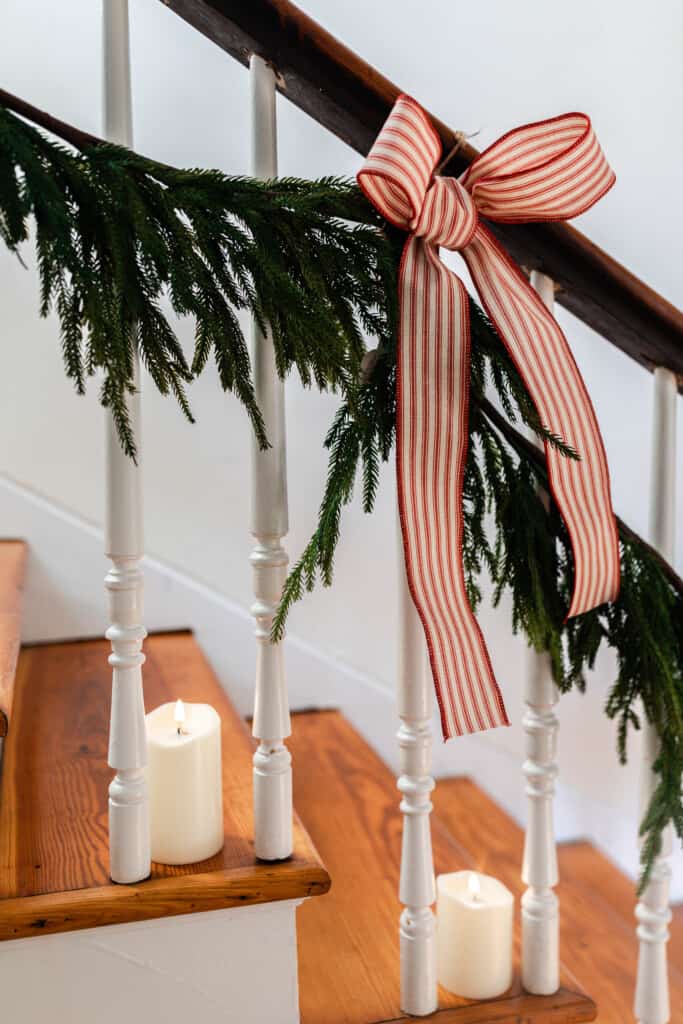 A close up picture of a stairway in a 1860's farmhouse line with greenery tied with red and white ticking stripe ribbon. Candles are placed on each stair.