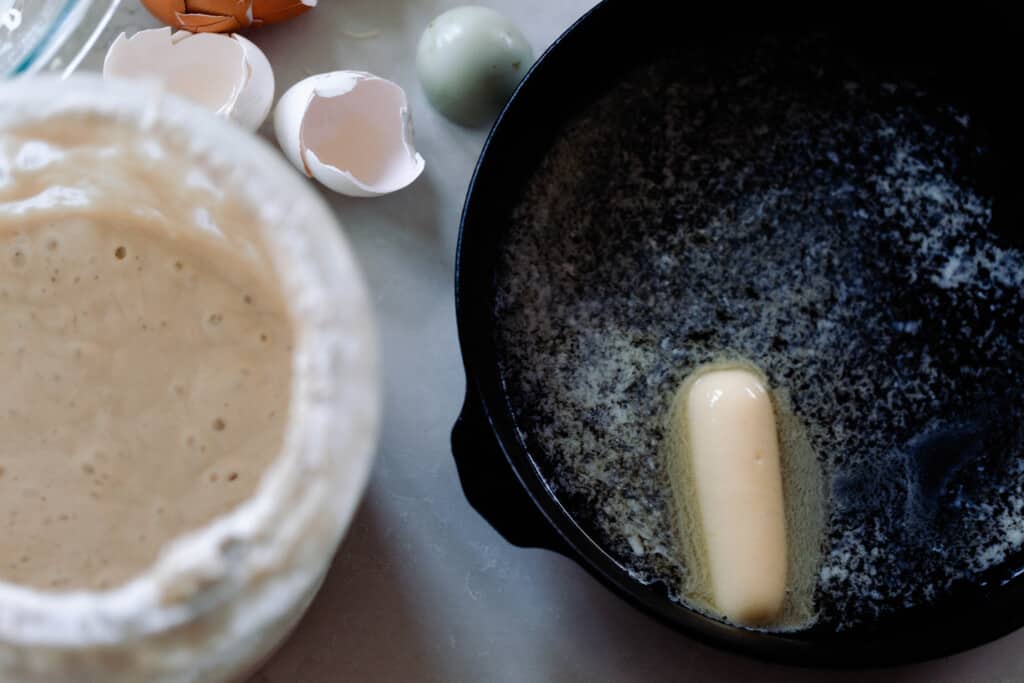 a stick of butter in a cast iron skillet melting. A glass container of sourdough starter sits to the left of the skillet