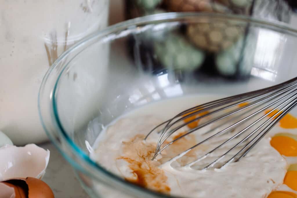 eggs, sourdough starter, vanilla, and milk in a glass bowl with the ingredients around the bowl on a white countertop