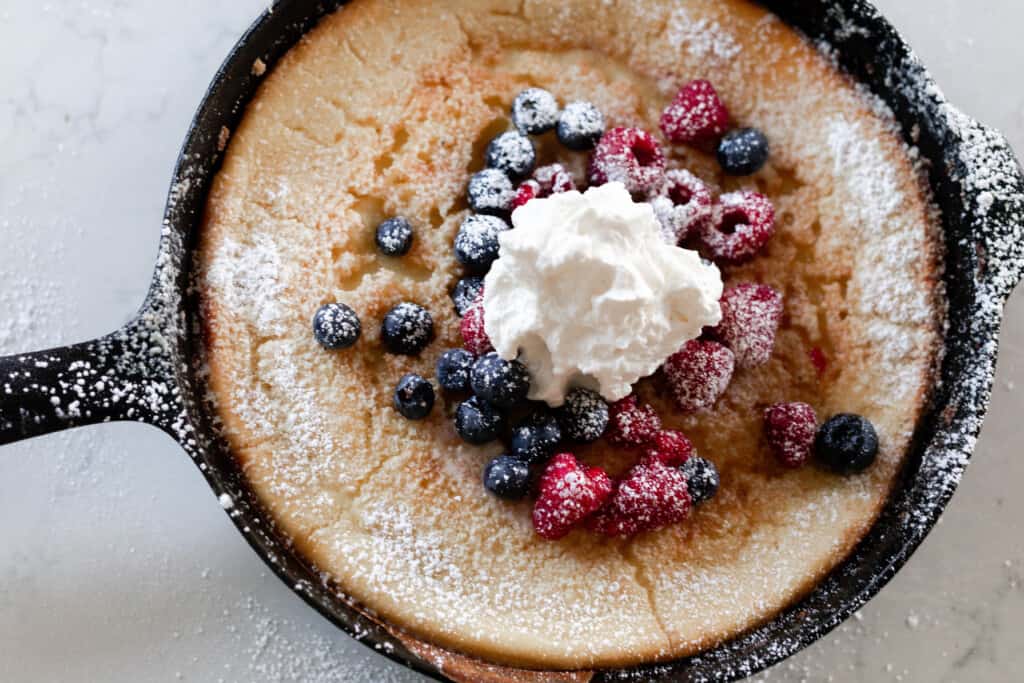 overhead photo of a cast iron skillet with a sourdough German pancake topped with berries, whipped cream, and powdered sugar