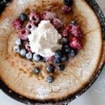 overhead photo of a cast iron skillet with a sourdough German pancake topped with berries, whipped cream, and powdered sugar