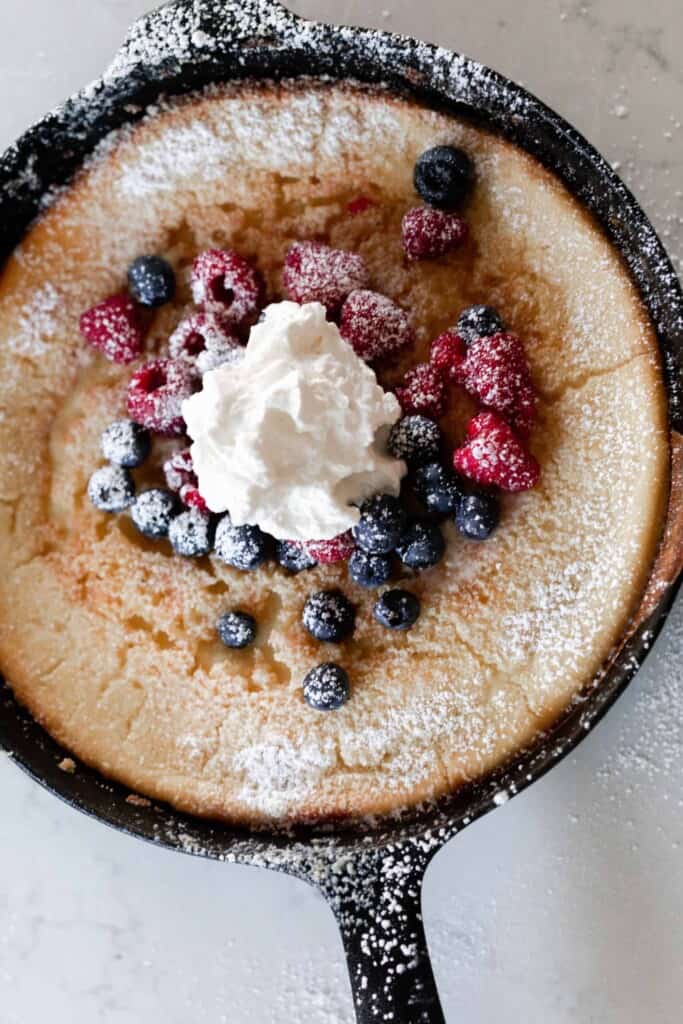 overhead photo of a cast iron skillet with a sourdough German pancake topped with berries, whipped cream, and powdered sugar