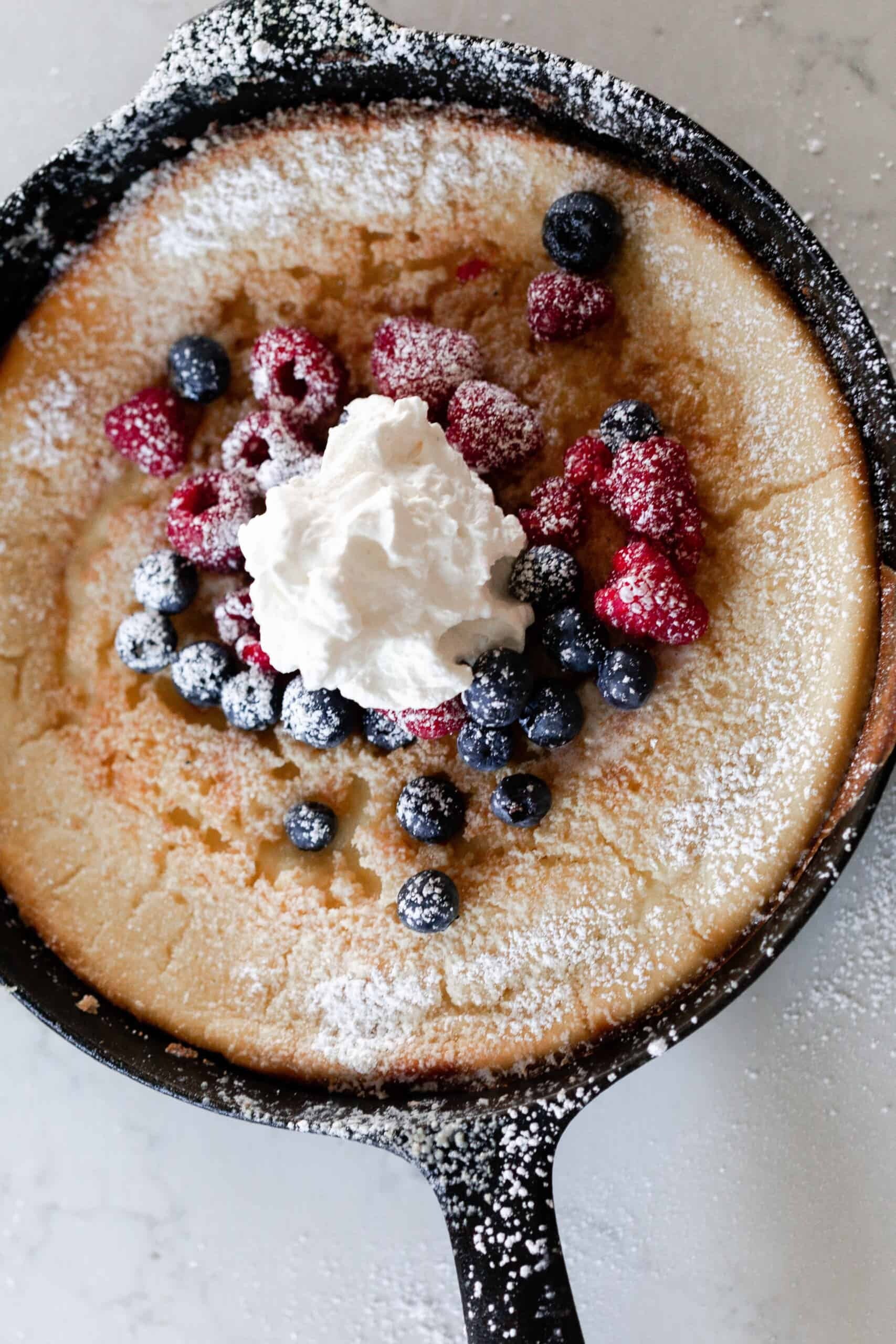 overhead photo of a cast iron skillet with a sourdough German pancake topped with berries, whipped cream, and powdered sugar