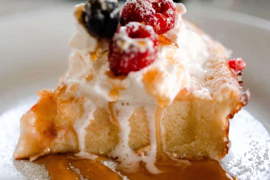 close up picture of a slice of sourdough dutch baby pancake stopped with fresh whipped cream, maple syrup, and fresh berries on a white plate