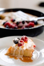 slice of sourdough dutch baby pancake topped with maple syrup, cream, and berries on a white plate with a cast iron skillet with more pancakes in the background