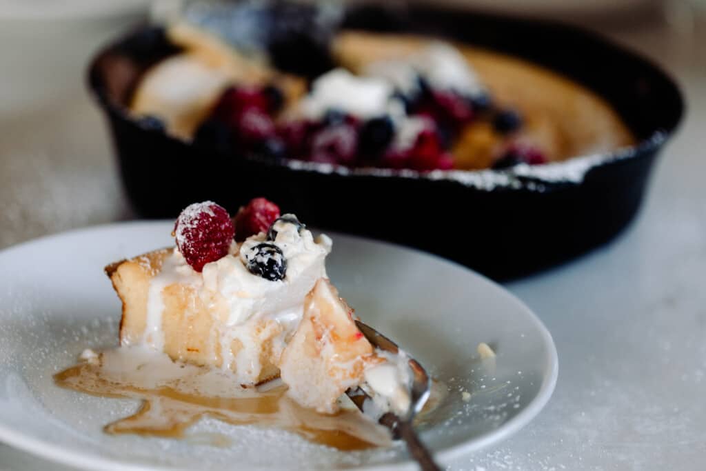 slice of sourdough German pancake topped with whipped cream, fresh berries, and maple syrup on a white plate with a cast irons skillet of more pancake and berries in the background