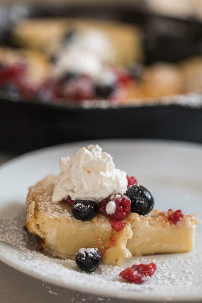slice of sourdough dutch baby pancake topped with whipped cream, fresh berries, and maple syrup on a white plate with a cast iron skillet with more pancakes and berries in the background