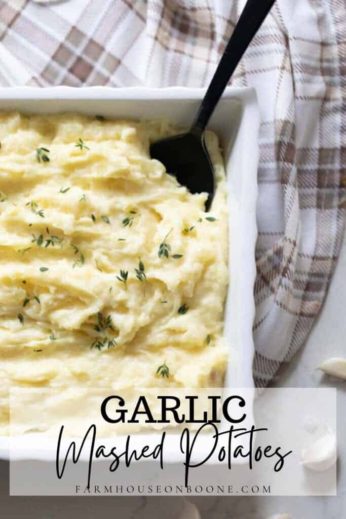 garlic mashed potatoes topped with herbs in a white baking dish with a black spoon in the potatoes. The baking dish rests on a tan and white plaid towel