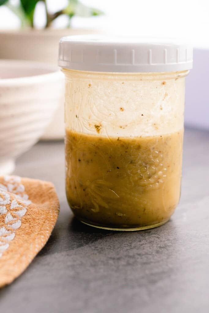 Greek marinate in a glass jar with a white lid on a gray countertop with a mustard towel to the left and a white bowl in the background