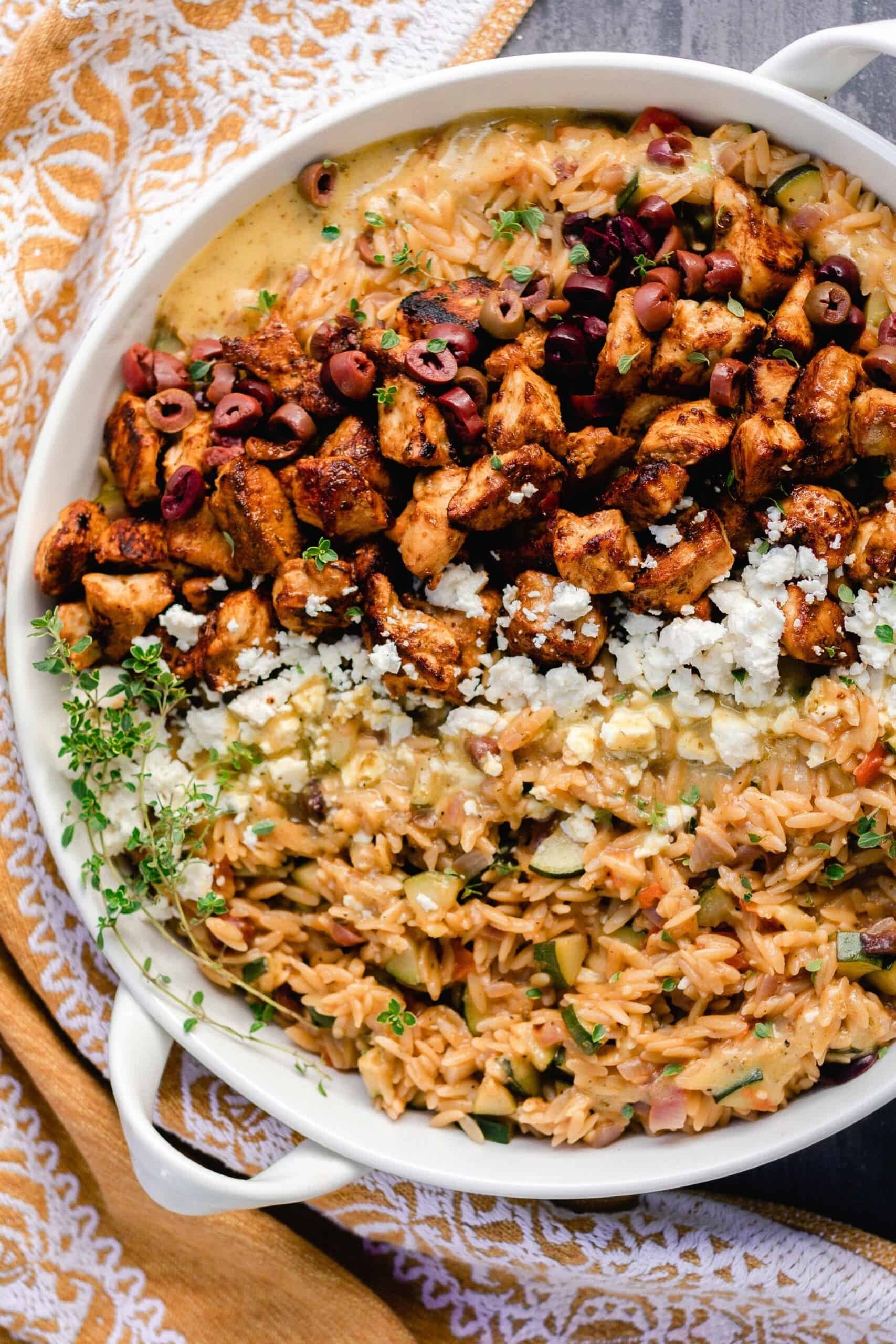 overhead photo of Greek chicken with orzo pasta, feta, and retables in a white enameled cast iron skillet on a gray countertop and a mustard colored towel to the left