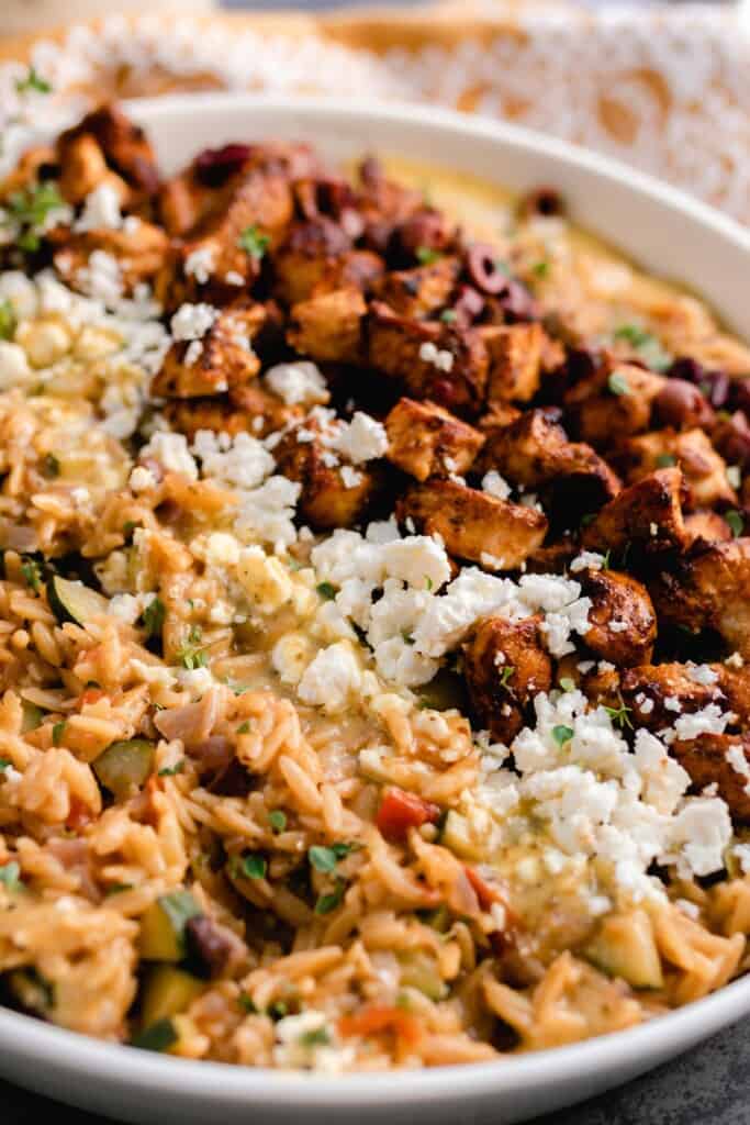 close up picture of a white dish of greek chicken with orzo pasta, feta cheese, and olives