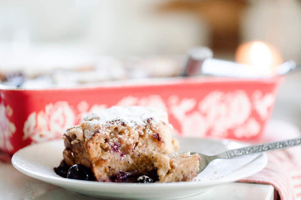 slice of stuffed sourdough French toast casserole on a white place with a fork taking out a bite of the casserole.  A red and white floral baking dish sits directly behind