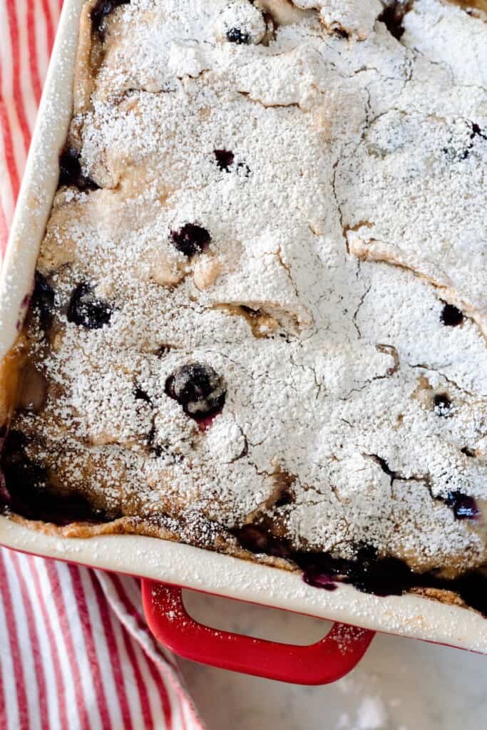 overhead photo of a baking dish with marscapone stuffed sourdough French toast casserole dusted with blueberries and dusted with powdered sugar in a red and white baking dish.