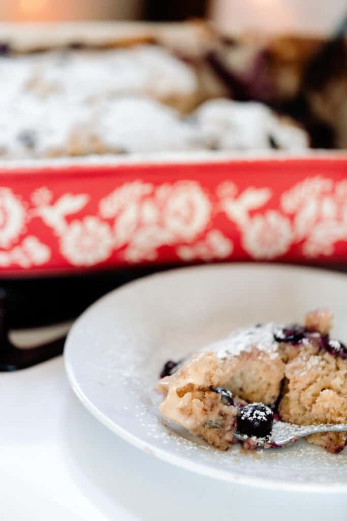 slice of marscapone stuffed sourdough French toast casserole with blueberries and dusted with powdered sugar on a white plate. A red and white casserole dish with the remaining casserole is directly behind the plate on the stove
