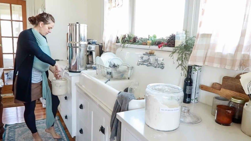 a woman in her white farmhouse kitchen holding her jar of sourdough starter under a Berkey water filter adding water to it