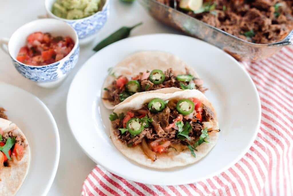 a large white plate with two tortillas topped with beef carnita meet, fermented jalapeños, cilantro, and tomatoes on a red and white stripped towel. Little white dishes with more taco toppings