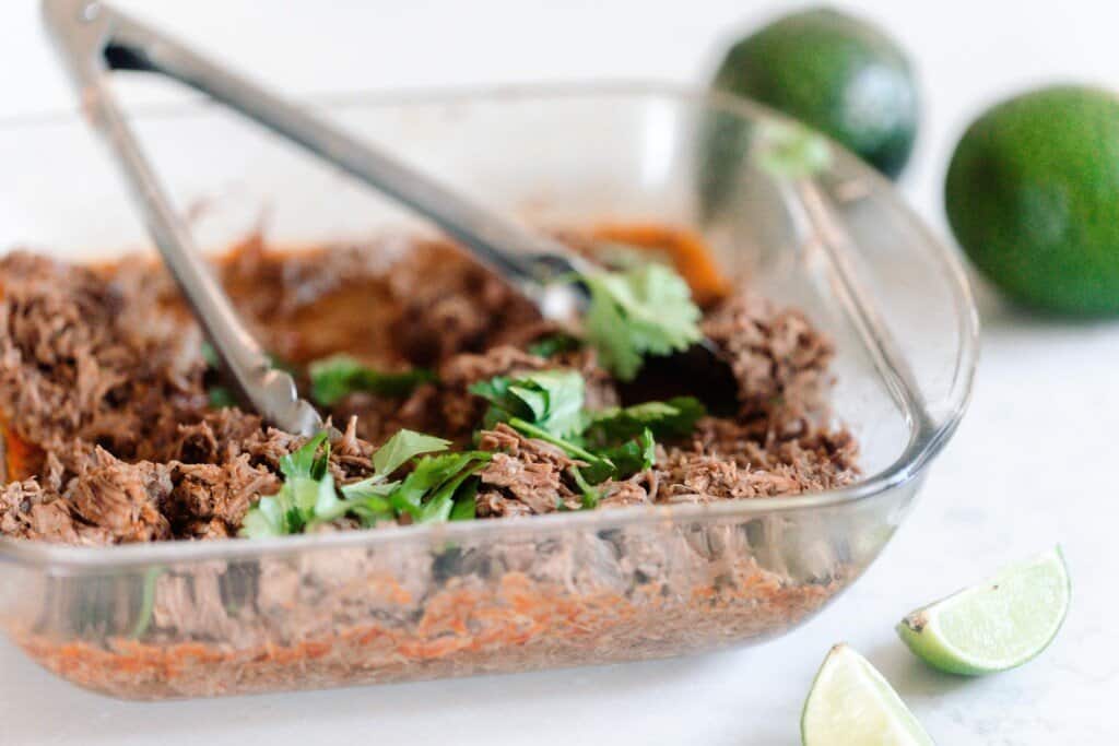 side view of a glass baking dish with beef carnitas topped with fresh cilantro on a white countertop, two limes are in the background