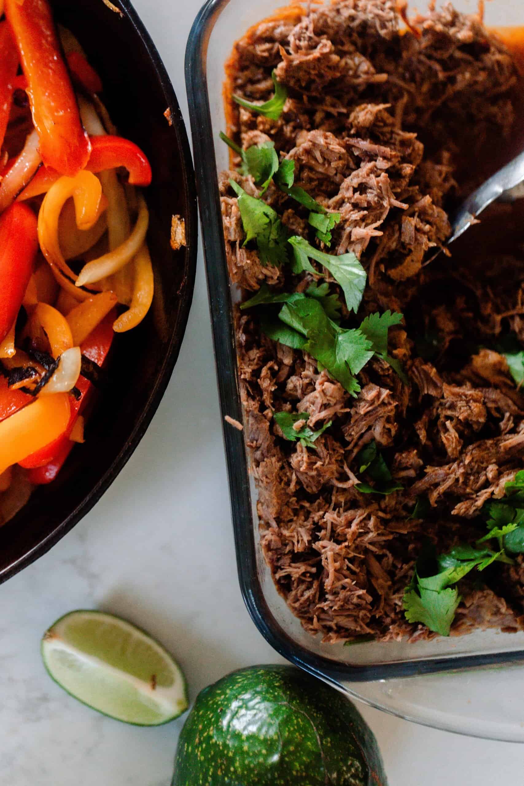 a glass dish of beef carnitas topped with cilantro. A cast iron skillet with peppers and onions are to the left and lime wedges are in front