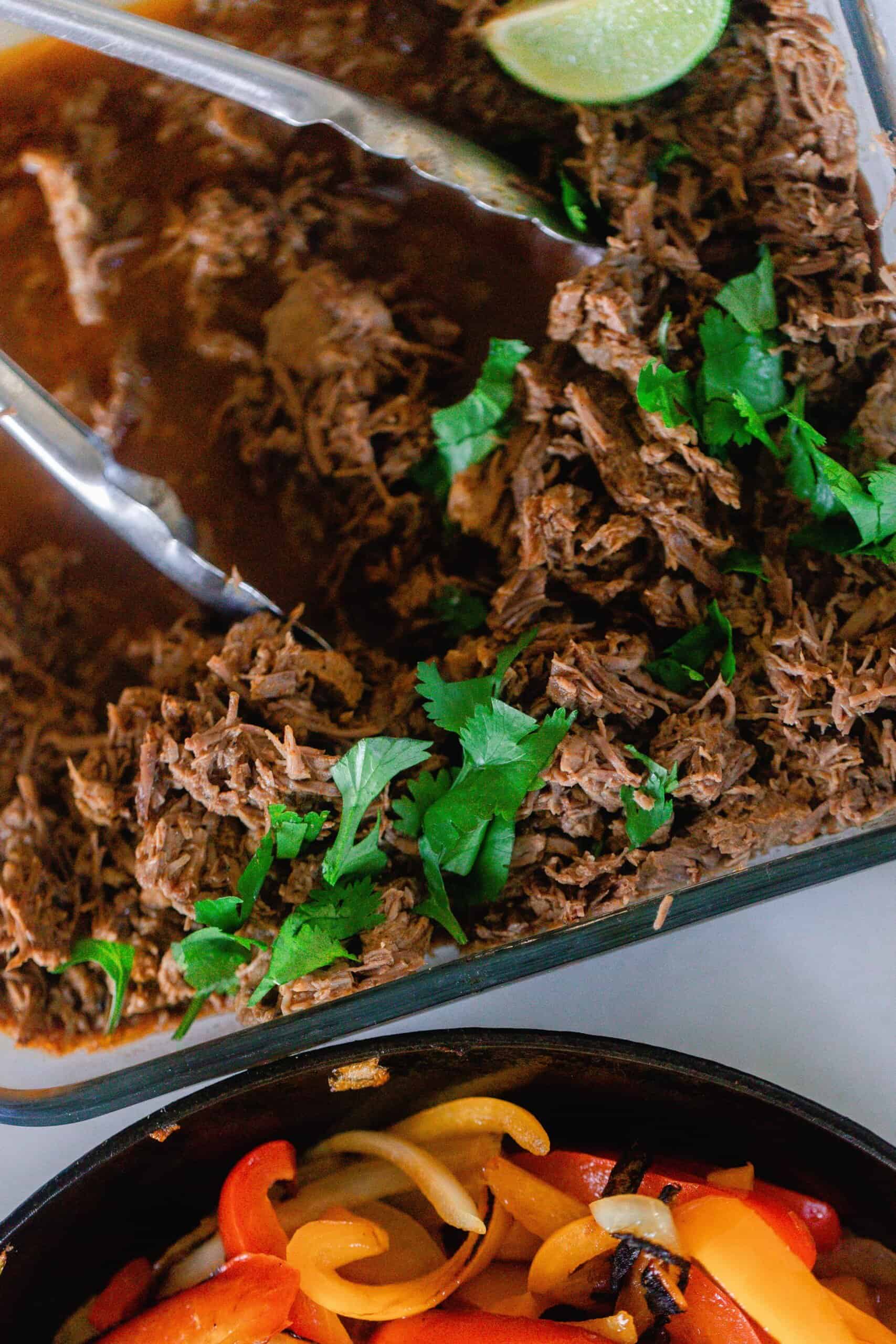 overhead photo of beef carnita meat topped with fresh cilantro in a glass backing dish with tongs and a cast iron skillet with cooked peppers and onions