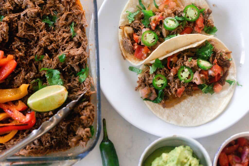 beef carnita tacos with toppings on a white plate next to a glass baking dish of carnita meat