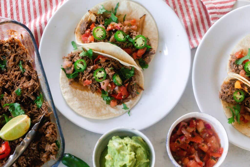 two beef carnitas served on a tortilla with jalapeños, and tomatoes on tortillas on a white plate with a dish of carnita meat to left and topping ingredient in white bowls in front