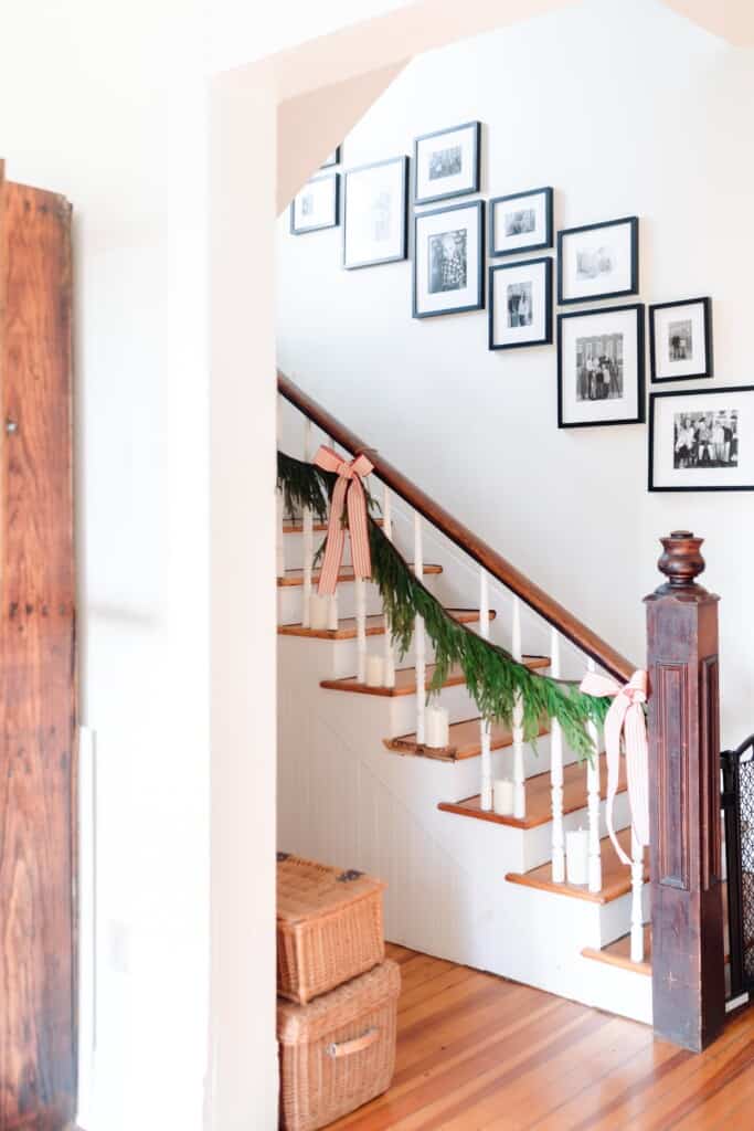 side view of a staircase gallery wall of black frames with family pictures are hung up the wall. The vintage staircase is decorated for Christmas with greenery, ribbons, and candles.