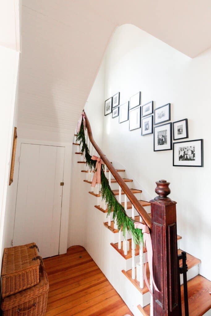 A vintage farmhouse stairway decorated for Christmas with garland, ribbon, and candles. A gallery wall of black and white family pictures in black picture frames goes up the wall. Two vintage baskets sit on the floor to the left of the stairway.