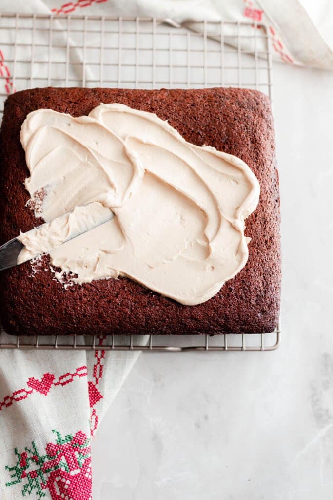 einkorn gingerbread cake with a little bit of mocha frosting being spread on top. The cake sits on a baking rack with a red and white towel underneath.