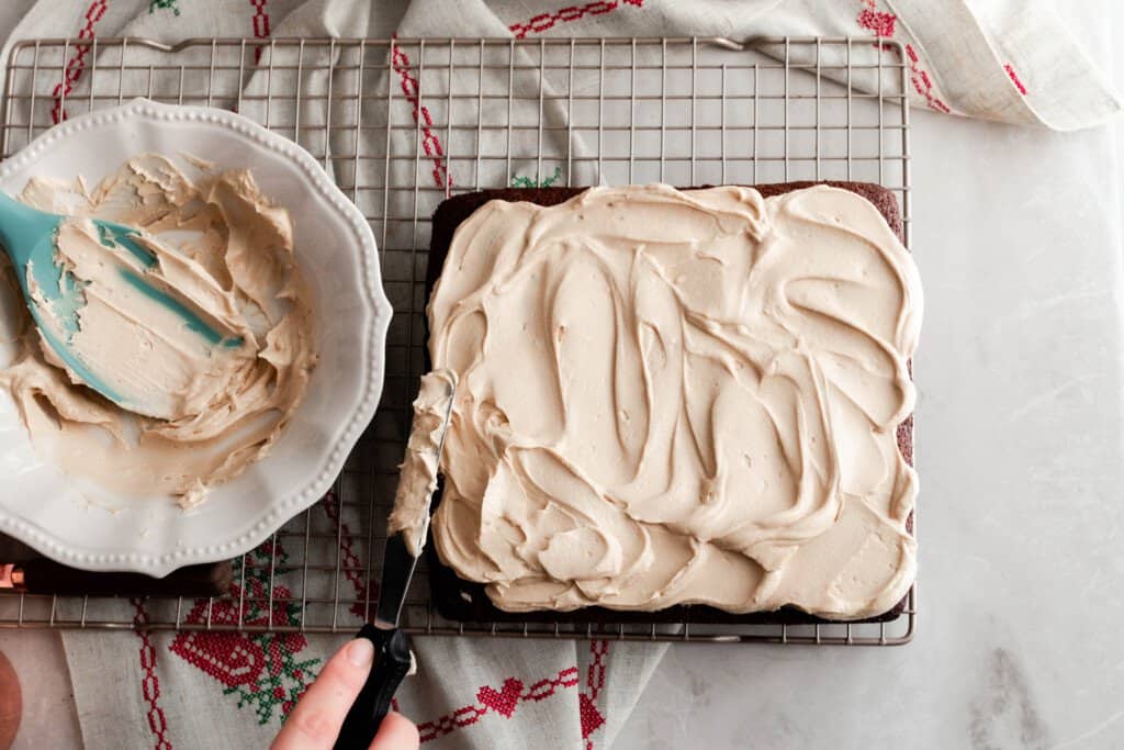 hand using a metal spatula to frost an einkorn cake with mocha frosting on a baking rack with a red and white towel underneath. To the left of the cake a bowl of frosting with a silicon spatula in the bowl