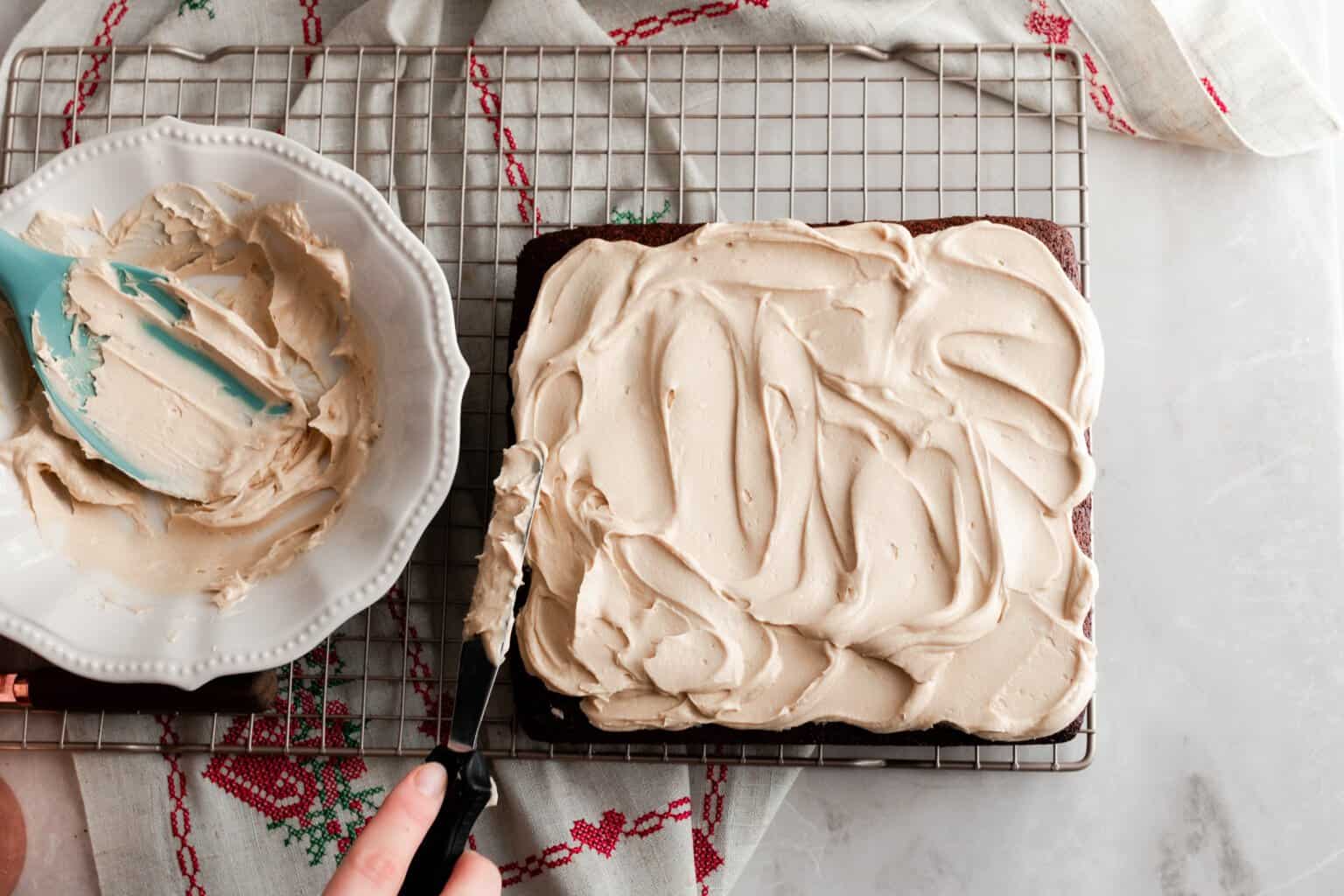 hand using a metal spatula to frost an einkorn cake with mocha frosting on a baking rack with a red and white towel underneath. To the left of the cake a bowl of frosting with a silicon spatula in the bowl