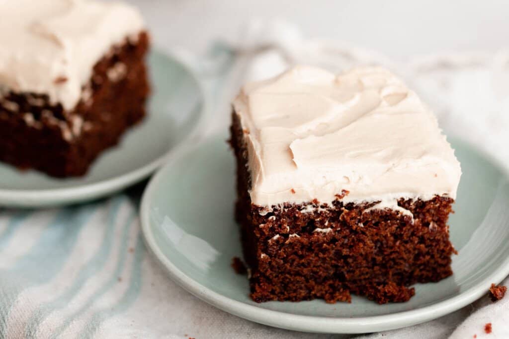 two square slices of einkorn gingerbread cake topped with thick layer of mocha frosting on light teal plates on a white and light blue stripped napkin