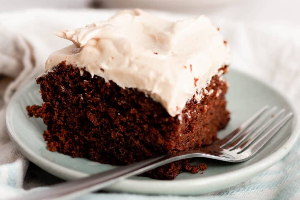close up picture of a large square slice of gingerbread einkorn cake with mocha frosting on a light teal plate with a fork resting on the plate
