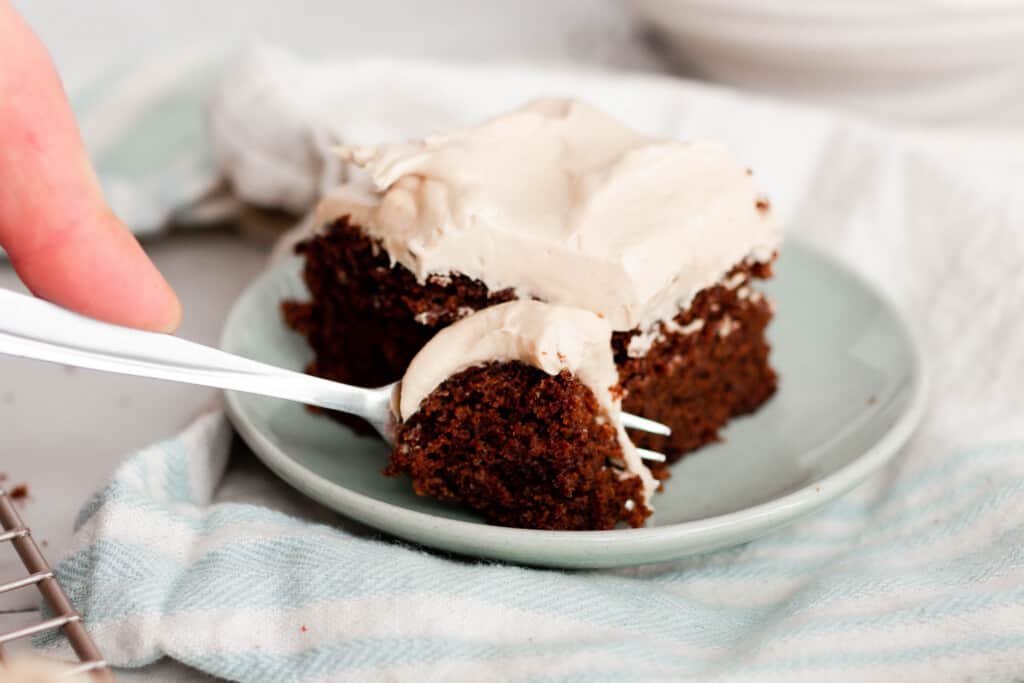 hand holding a fork taking out a forkful of gingerbread cake with mocha frosting on a white plate on top of a white napkin.