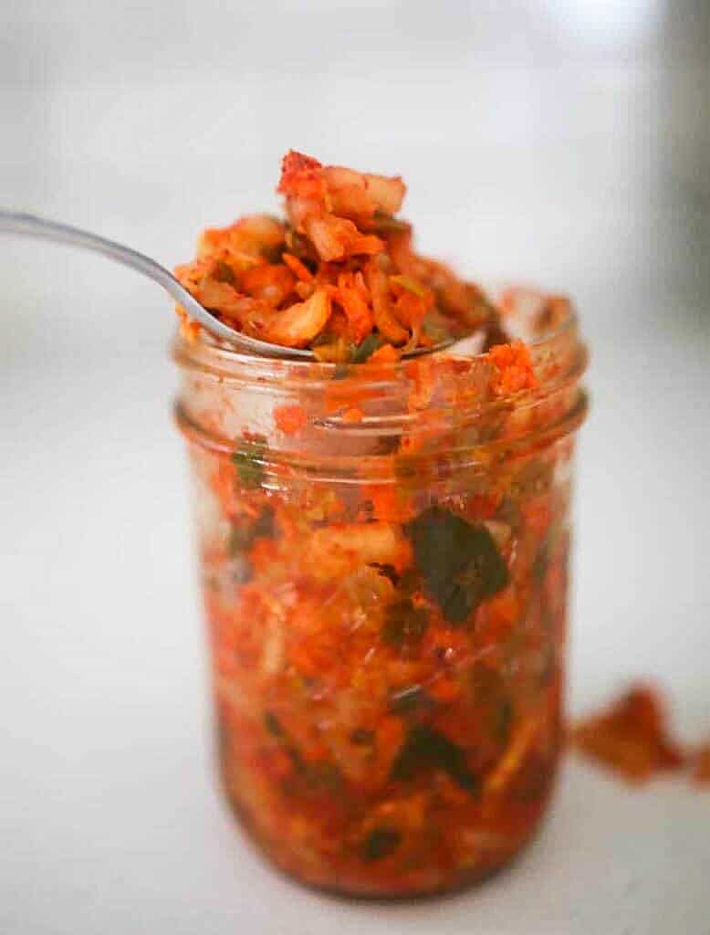 a spoonful of kimchi is coming out of a glass mason jar of kimchi that is sitting on a white countertop