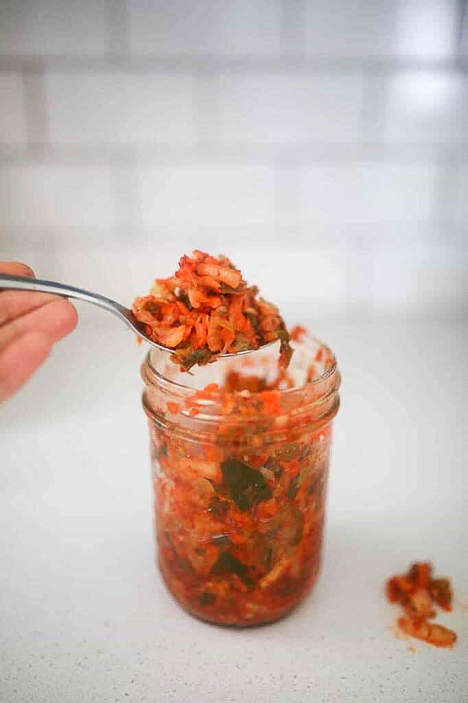 spoonful of homemade kimchi coming out of a mason jar on a white countertop with a white subway tile backsplash