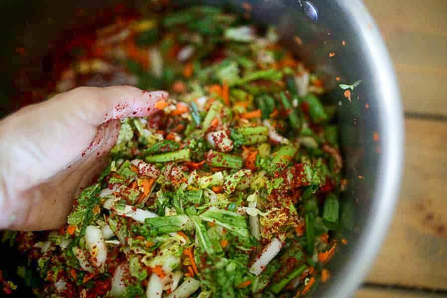 a hand in a metal bowl holding cabbage and making homemade kimchi with Napa cabbage, Korean chili flakes, carrots in a metal bowl.