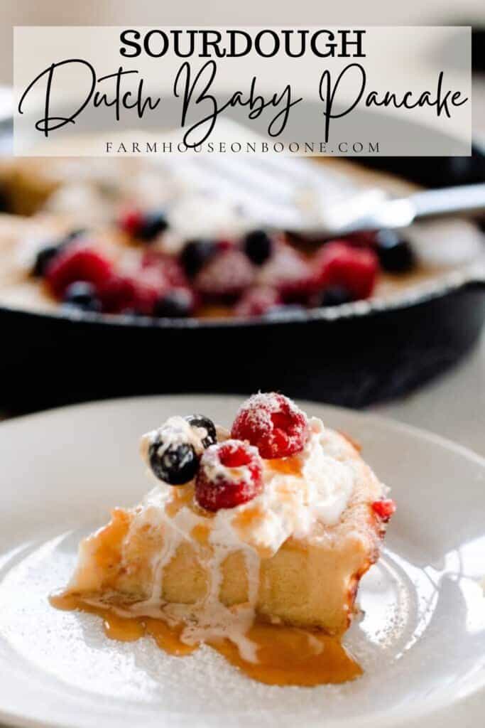 slice of sourdough dutch baby pancake topped with maple syrup, cream, and berries on a white plate with a cast iron skillet with more pancakes in the background