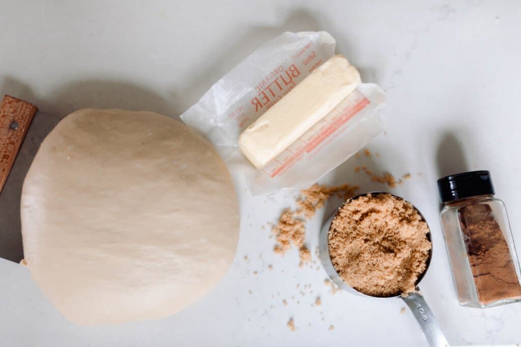 sourdough cinnamon roll dough in a ball on a white countertop. a stick of butter, brown sugar in a measuring cup and cinnamon in a container sit to the right of the dough
