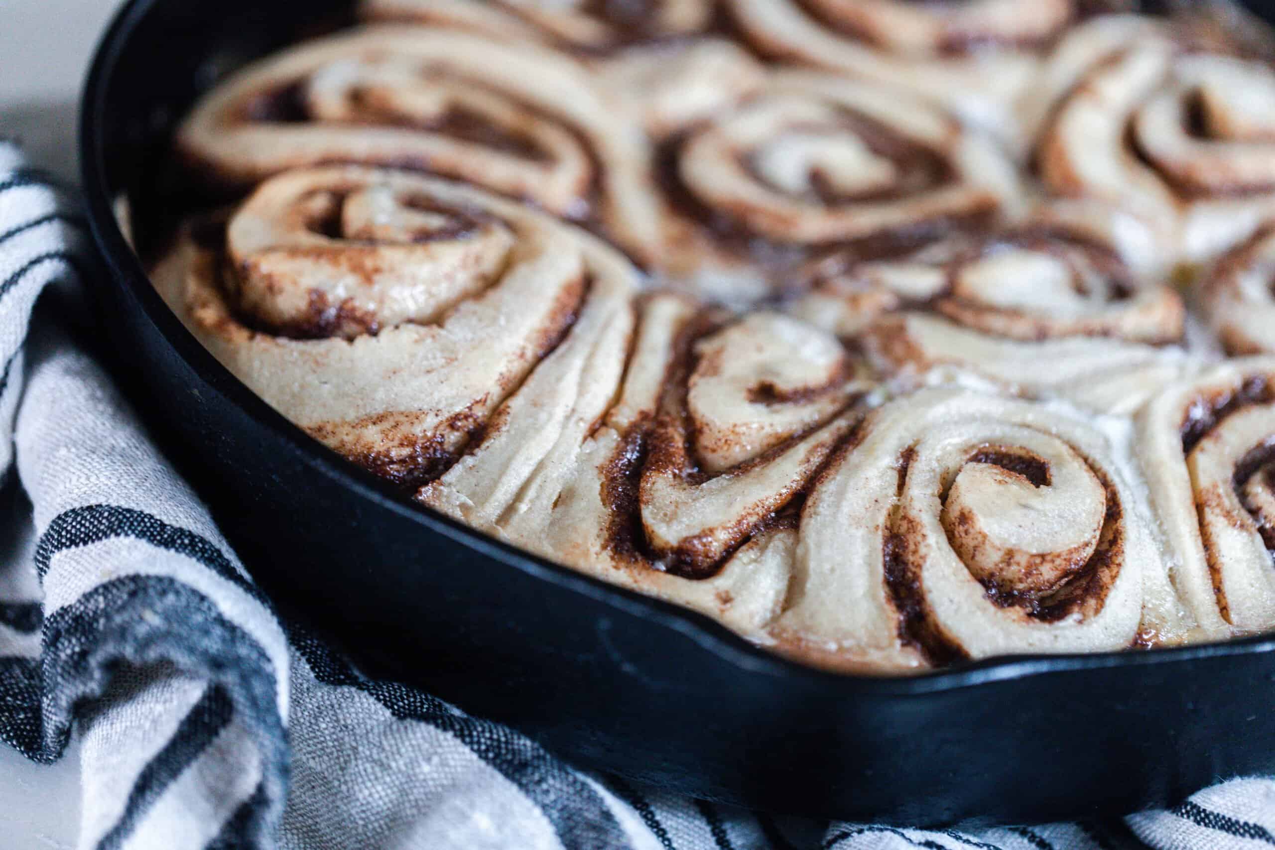 sourdough cinnamon buns fresh out of the oven in a cast iron skillet on top of a white and black stripped towel