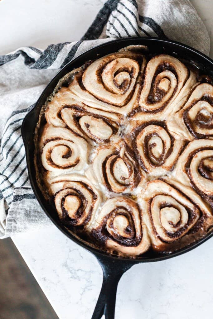 overhead photo of sourdough cinnamon rolls in a cast iron skillet on top of a white and black stripped towel on a white quartz countertop