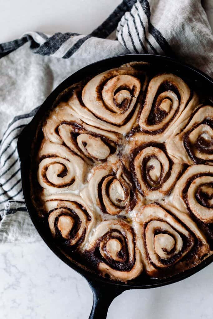overhead photo of sourdough cinnamon rolls in a cast iron skillet on a white and black stripped towel on a white quartz countertop