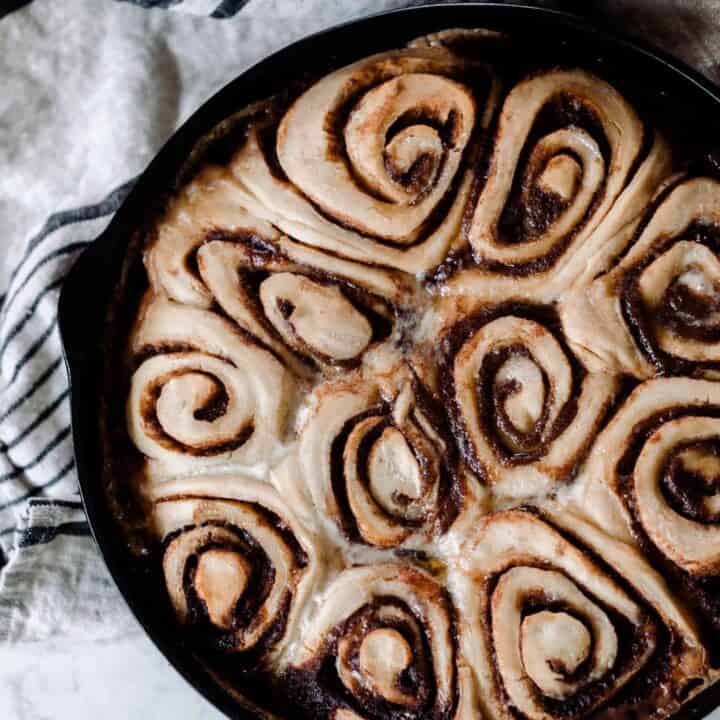 overhead photo of sourdough cinnamon rolls in a cast iron skillet on a white and black stripped towel on a white quartz countertop