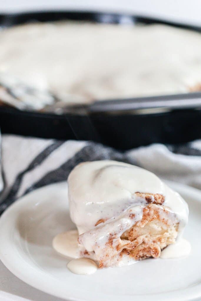 sourdough cinnamon bun with cream cheese icing on a white plate with a cast iron skillet of more cinnamon rolls on top of a white and black stripped towel