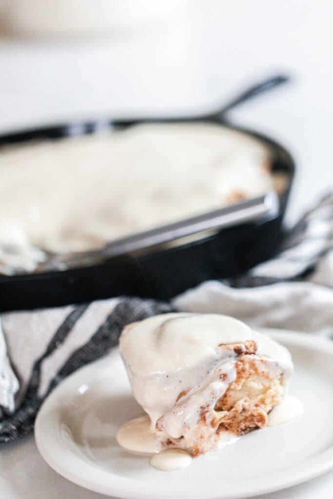 sourdough cinnamon roll covered in cream cheese icing on a white plate with a cast iron skillet full of rolls in the background