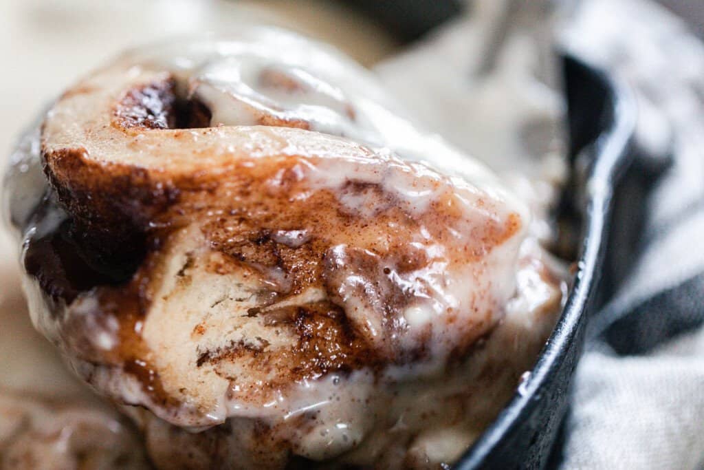 close up side view of a fluffy sourdough cinnamon roll with icing being picked up out of a cast iron skillet with a spatula