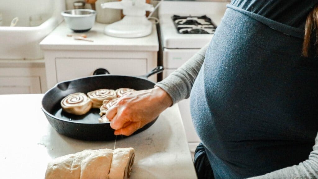 woman wearing a baby in a blue wrap is using string to slice sourdough cinnamon rolls and place them in a cast iron skillet